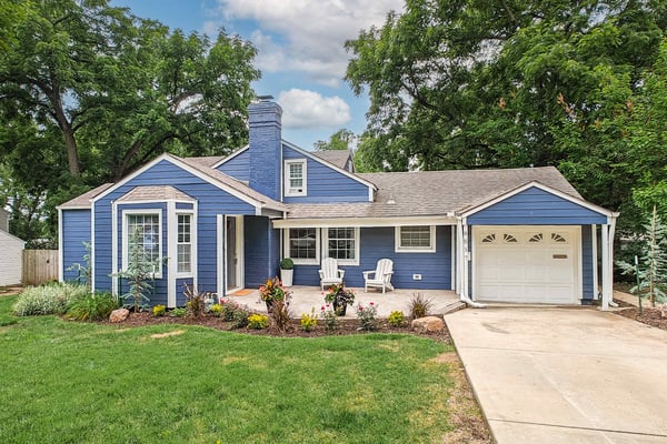 Blue cottage-style house with attached garage, front patio with Adirondack chairs, landscaped lawn and garden beds surrounded by mature trees.